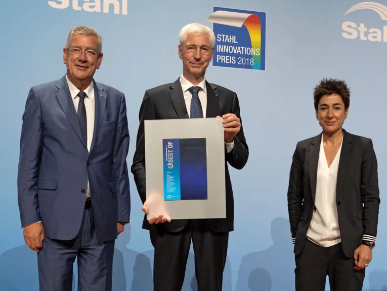 Zwei Männer und eine Frau, ein Mann hält eine Urkunde in die Kamera.